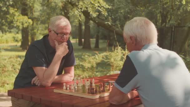 Taille haute de deux amis aînés caucasiens assis devant l'autre à la table dans le parc en jouant aux échecs - Séquence, vidéo