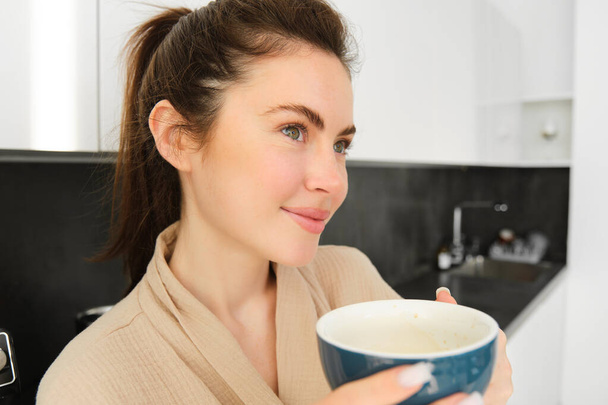 Πορτρέτο της όμορφης νεαρής γυναίκας με μπουρνούζι, πίνοντας πρωινό καφέ και απολαμβάνοντας τη γεύση, χαμογελώντας ευχαριστημένος, στέκεται στην κουζίνα. - Φωτογραφία, εικόνα