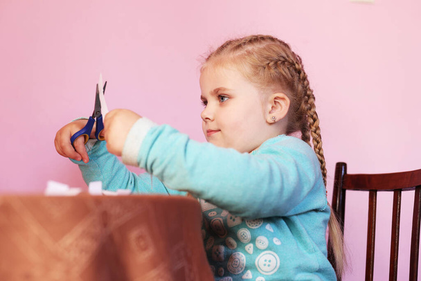 A child cuts paper with scissors. A little girl is sitting and cutting white paper. A preschooler learns to work with scissors. Program of preschool education at home. Child development, creativity - Photo, Image