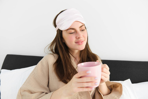 Mulher linda em pijama e máscara de dormir, segurando xícara de café, bebendo cappuccino em sua cama e olhando feliz, descansando no quarto no fim de semana, tendo manhã relaxante. - Foto, Imagem