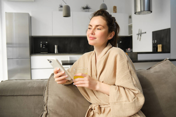 Piękna młoda kobieta odpoczywa na kanapie, ciesząc się porankiem w domu, trzymając szklankę soku pomarańczowego i smartfona, patrząc przez okno. - Zdjęcie, obraz