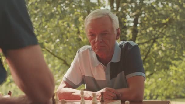 Vyötärö ylös vanhempi valkoihoinen mies etsii pensiivisesti shakkilauta pelatessaan mies ystävä ulkona puistossa - Materiaali, video