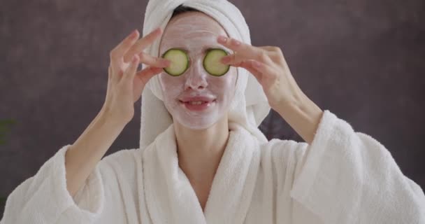 Fröhliche Frau in Bademantel und Handtuch auf dem Kopf mit Gesichtsmaske für die Reinigung und gesunde Haut, Spaß haben, lächeln und die Augen mit Gurkenscheiben bedecken. Hautpflege und Selfcare-Konzept - Filmmaterial, Video