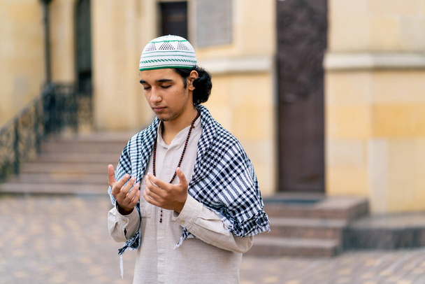 Un chico de aspecto árabe con ropas nacionales con un rosario alrededor de su cuello reza en el patio de la mezquita - Foto, Imagen