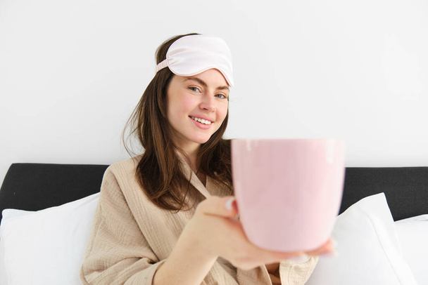 Piękna uśmiechnięta nowoczesna kobieta w piżamie i masce do spania, oferująca filiżankę herbaty lub kawy w łóżku i wyglądająca na szczęśliwą, siedzącą w sypialni i relaksującą. - Zdjęcie, obraz