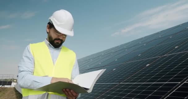 ソーラーパネルを検査する男性エンジニア. 太陽エネルギーの取付けのパネルを点検して下さい. 太陽電池は重要な再生可能エネルギーになるだろう. 制服の技術者はノートにノートを作ります. - 映像、動画