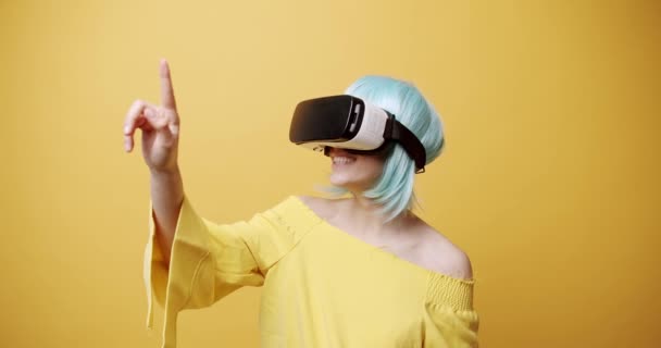 Mujer asombrada explorando la realidad virtual. Foto estática de una joven sorprendida con pelo azul y casco VR mirando a su alrededor y tocando objetos invisibles sobre fondo amarillo - Imágenes, Vídeo