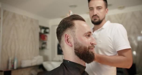 Attractive male is getting a modern haircut in barber shop. Barber sets hair by spray and combs them. Barbershop. Hairdresser doing haircut in retro hair salon. Mens hair styling. - Footage, Video
