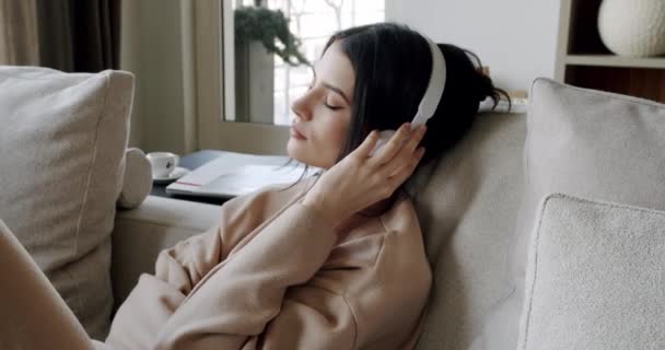 Mindful young happy woman wearing wireless headphones, listening relaxing music, resting on comfortable sofa, enjoying interesting audio book or affirmations, spending leisure indoors - Footage, Video