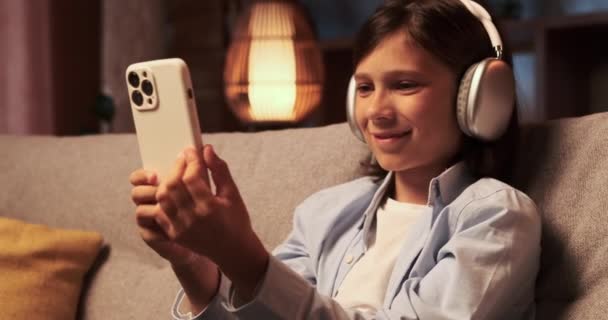 Boy wearing headphones sits with a smile, engrossed in phone. His eyes reflect amusement, and lips curve into a cheerful grin as he interacts with the device. - Footage, Video