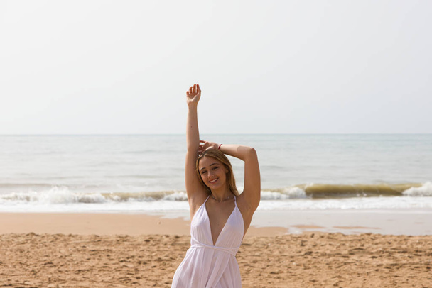 Νεαρή όμορφη ξανθιά γυναίκα σε λευκό φόρεμα είναι το περπάτημα στην άμμο στην ακτή της παραλίας σε μια ηλιόλουστη μέρα. Η γυναίκα κάνει διαφορετικές εκφράσεις του σώματος. Στο βάθος η γαλάζια θάλασσα. - Φωτογραφία, εικόνα