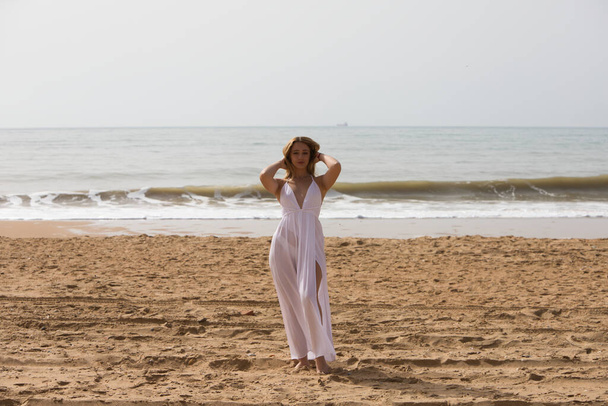 Jeune belle femme blonde en robe blanche marche sur le sable sur le rivage de la plage par une journée ensoleillée. La femme fait différentes expressions corporelles. En arrière-plan la mer bleue. - Photo, image