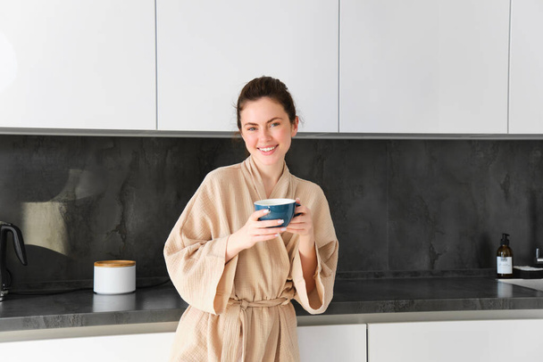 Portrait de jeune femme heureuse commence sa matinée avec une tasse de café, buvant du thé dans une tasse, debout dans la cuisine, souriant joyeusement. - Photo, image