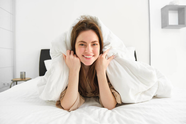 Cute girl with messy hair, lying in bed covered in white sheets duvet, smiling and laughing coquettish, spending time in her bedroom. - Photo, Image