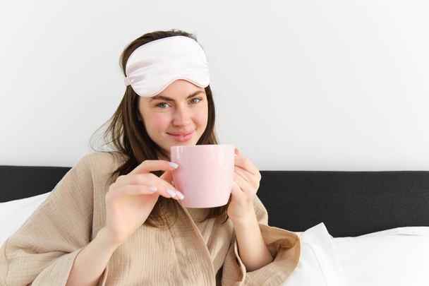Ευτυχισμένη νεαρή γυναίκα που ξεκινά το πρωί της με ένα φλιτζάνι καφέ στο κρεβάτι, χαμογελώντας και απολαμβάνοντας τεμπέλικη μέρα της, έχει μάσκα ύπνου στο μέτωπο. - Φωτογραφία, εικόνα