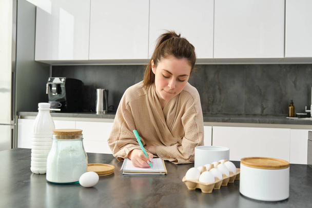 Vonzó fiatal vidám lány süt a konyhában, tésztát csinál, receptkönyvet tart, ötletei vannak. - Fotó, kép