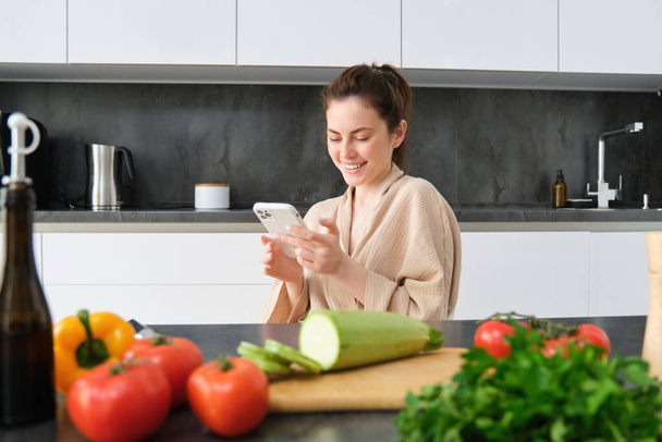 スマートフォンでキッチンに座っているバスローブの女性の肖像画,夕食を調理,ソーシャルメディアでレシピを見る,食事の準備方法のビデオチュートリアル. - 写真・画像