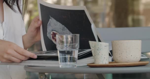 Widok upraw na młodą kobietę siedzącą przy stole ulicznej kawiarni, przewracając książkę i oglądając zdjęcia podczas przerwy na śniadanie i kawę. - Materiał filmowy, wideo