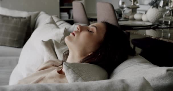 Calm young woman having healthy daytime, relaxing on couch with eyes closed and hands behind head, peaceful girl sleeping breathing fresh air resting leaning on comfortable sofa at modern home. - Filmmaterial, Video