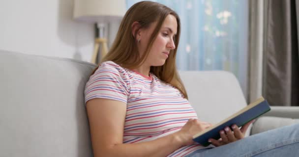 Mladá hezká žena čte knihu, zatímco odpočívá na pohodlné pohovce doma. Vysoce kvalitní 4K záběry - Záběry, video