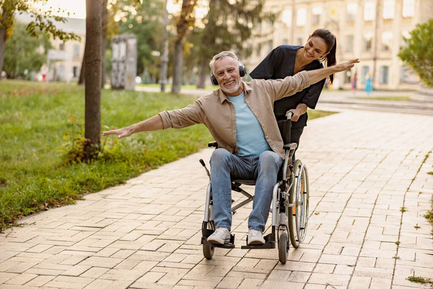 Joyeux homme handicapé mature en fauteuil roulant portant des écouteurs s'amuser lors d'une promenade dans la ville assisté par une belle jeune infirmière. Assistance, concept de réhabilitation - Photo, image