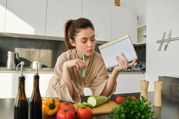 Porträt einer Frau, die ihre Einkaufsliste überprüft, Gemüse betrachtet, ein Notizbuch in der Hand hält, während sie in der Küche das Rezept liest, Tomaten und Zucchini hackt. - Foto, Bild