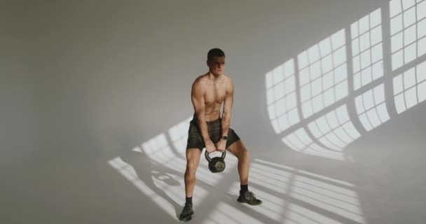 Atletický mladý muž dělá dřepy cvičení s kettlebell v rukou ve studiu s bílým pozadím. Samec bez košile se svalnatým tělem má cvičení s tělesnou hmotností. - Záběry, video
