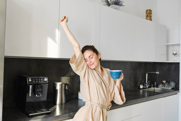 Portrait of happy girl dancing with coffee in the kitchen, wearing bathrobe, enjoying her morning routine. - Photo, Image