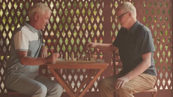 Mittleres Porträt zweier kaukasischer älterer männlicher Freunde, die für die Kamera posieren, während sie draußen am Tisch Schachspiel spielen - Filmmaterial, Video