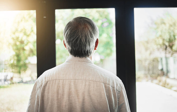 Back, mature man and thinking by window, depression and anxiety alone in home. Rear view, retirement and sad person contemplating in house, planning future and remember memory, idea or reflection. - Photo, Image