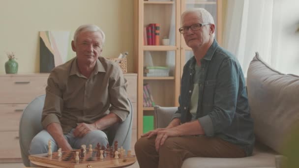 Μέτριο αργό πορτραίτο δύο Καυκάσιων ανδρών ηλικιωμένων φίλων που ποζάρουν για την κάμερα καθισμένοι ο ένας μπροστά στον άλλο στο ξύλινο τραπέζι παίζοντας σκάκι - Πλάνα, βίντεο