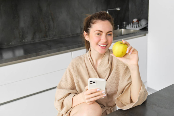 Detailní portrét usměvavé brunetky v županu, sedí doma v kuchyni, používá mobilní telefon a drží jablko, objednává čerstvé ovoce v aplikaci smartphone, nakupuje potraviny online, vyhledává recept. - Fotografie, Obrázek