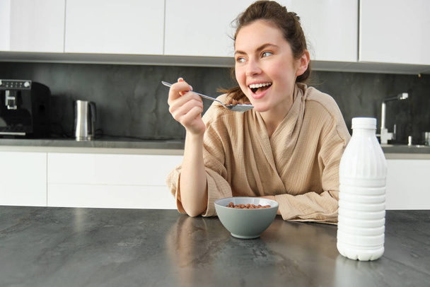 Portret van jonge mooie vrouw in badjas, ontbijtgranen etend, leunt op keukenblad, kijkend naar haar ochtendmaaltijd. - Foto, afbeelding