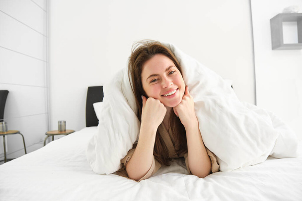 Portrait of gorgeous smiling woman, lying in bed covered in duvet, has messy hair, looking happy, relaxing in her bedroom, spending time in hotel room in morning. - Photo, Image