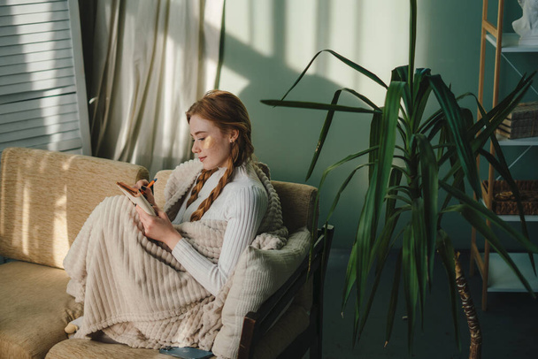 Piękna kobieta siedząca na kanapie w przytulnym swetrze i pisząca w notatniku. Koncepcja wakacji i wypoczynku - młoda kobieta z ołówkiem pisząca do pamiętnika w przytulnym domu - Zdjęcie, obraz