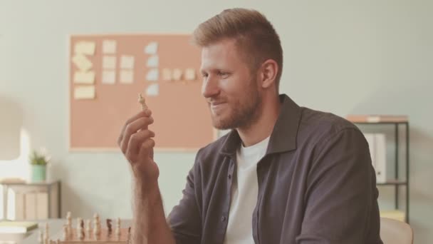 Medium slow mo portret van jonge blanke man met wit schaakstuk glimlachend op camera zittend door schaakbord aan tafel binnen - Video