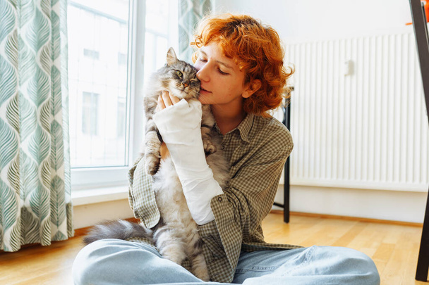 teen girl with curly red hair, broken arm in cast, sits on floor of house, hugging favorite fluffy cat. Friendship teenager pet - Фото, изображение