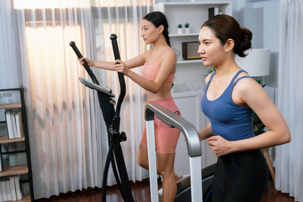 Mujer asiática atlética enérgica y fuerte corriendo en máquina elíptica en casa con compañero de entrenamiento o entrenador. Búsqueda de físico en forma y compromiso con un estilo de vida saludable. Vigoroso - Foto, Imagen
