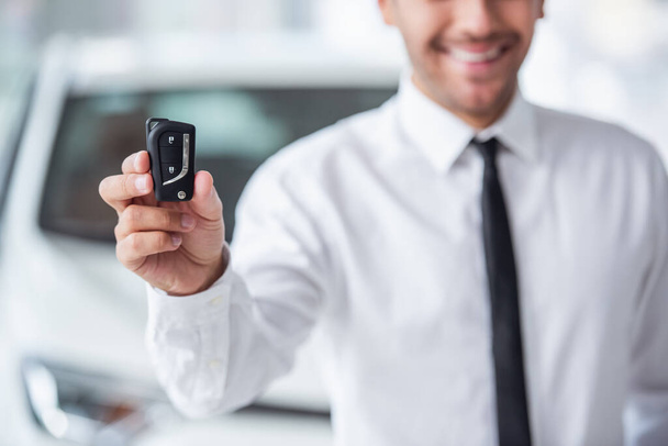 Imagen recortada de hombre de negocios guapo en traje sosteniendo la llave y sonriendo mientras está de pie en concesionario de automóviles
 - Foto, Imagen