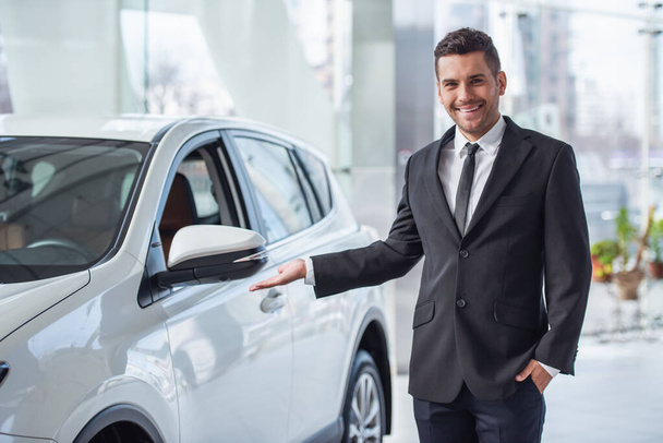 schöner Geschäftsmann im Anzug blickt in die Kamera und lächelt, während er neben dem Auto im Autohaus steht - Foto, Bild