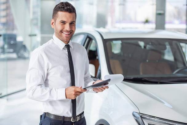 Красивый работник автосалона в костюме держит папку, смотрит в камеру и улыбается, стоя рядом с машиной
 - Фото, изображение