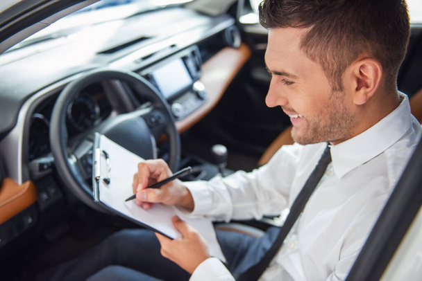 Красивый бизнесмен в костюме подписывает документы и улыбается, сидя в новой машине в автосалоне
 - Фото, изображение