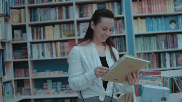 Mosolygó fiatal nő tölti szabadidejét a könyvesboltban, keres egy új könyvet vásárolni - Felvétel, videó
