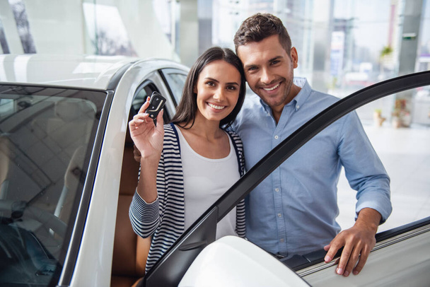 訪問車の販売代理店。美しいカップルは新しい車の鍵を持っていてカメラを見て笑っています - 写真・画像