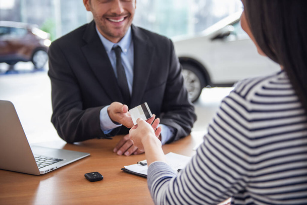 Visitando concesionario de coches. Guapo gerente de ventas está tomando una tarjeta de crédito del cliente y sonriendo
 - Foto, imagen