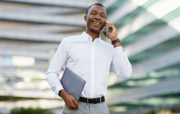 Деловой звонок. Улыбающийся черный бизнесмен разговаривает по мобильному телефону во время прогулки на свежем воздухе рядом с современным офисным центром, симпатичный афроамериканский предприниматель держит ноутбук, наслаждаясь приятной беседой - Фото, изображение