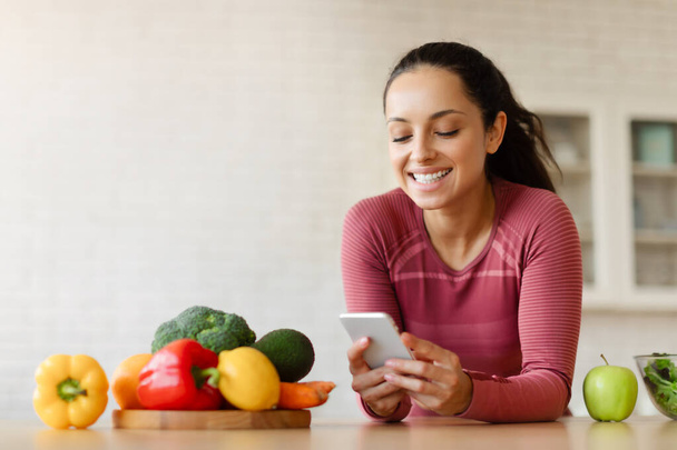 スリミングアプリ。 現代のキッチンインドアで携帯電話の調理健康的な食事,スマートフォンとウェブサーフィンでポジショニングで減量アプリケーションを使用してスポーツウェアの若いフィットレディ - 写真・画像