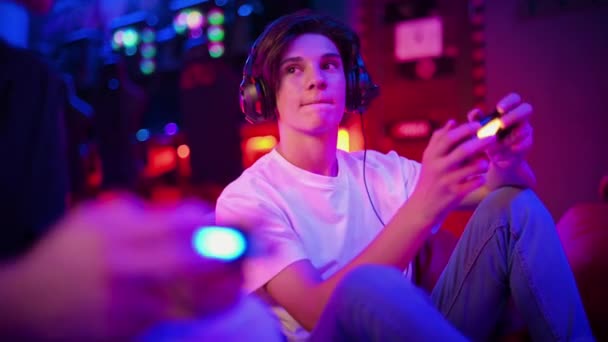 Due amici adolescenti in cuffia stanno parlando e giocando a una console di gioco usando gamepad, parlando e ridendo mentre sono seduti su borse di fagioli. Illuminazione blu e rossa - Filmati, video