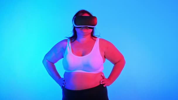 Женщина с избыточным весом в спортивном костюме и VR гарнитуре позирует в студии с синим фоном и красной подсветкой, глядя в камеру - Кадры, видео