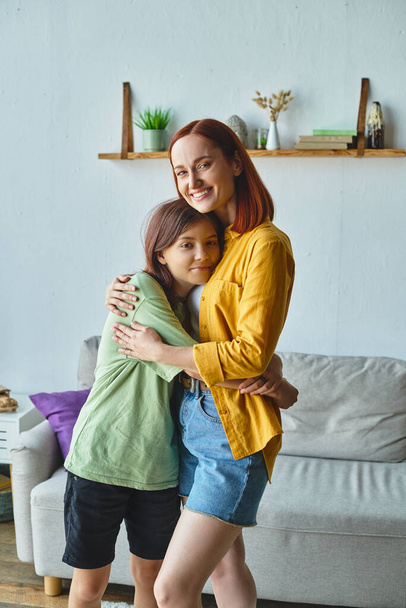 mujer alegre con hija adolescente abrazando y mirando a la cámara en casa en acogedora sala de estar - Foto, imagen